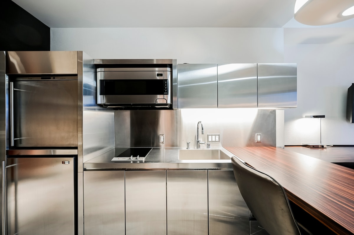 The kitchenette features modern stainless steel appliances, including a refrigerator, microwave, and flat top stove. The sleek cabinets provide ample storage, and the countertop offers space for meal preparation. A dining area is visible, equipped with a stylish chair and wooden table.