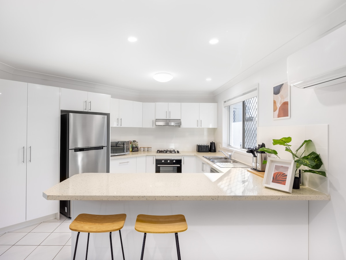 A modern kitchen is equipped with sleek white cabinetry and quality appliances, including a refrigerator and oven. A countertop with two wooden bar stools overlooks the cooking area. Natural light enters through a nearby window, accentuating the clean lines and minimalist decor.