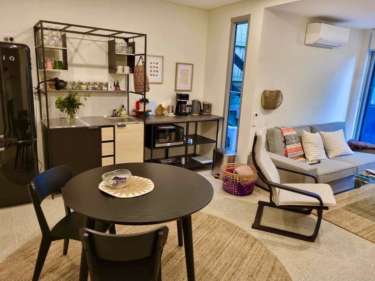 A modern kitchenette and living area feature a round dining table, accompanied by four chairs. A well-stocked shelf and appliance unit are visible, showcasing kitchen essentials. A comfortable sofa is positioned nearby, enhancing the cozy ambiance. Natural light streams through large windows.