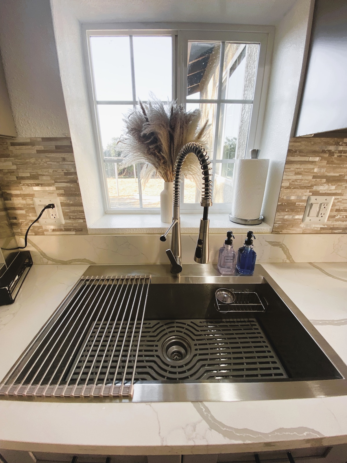 A modern kitchen sink is placed under a window, showcasing a sleek stainless-steel design. A decorative arrangement of dried plants is positioned nearby, with a roll of paper towels and two bottles of liquid soap visible on the countertop.