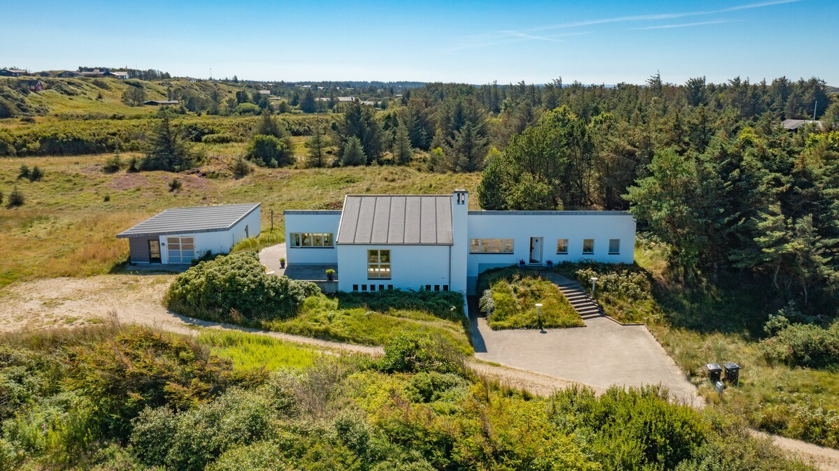 A modern villa is situated amidst lush greenery and natural landscapes. The streamlined structure features large windows and a flat roof, with pathways leading up to the entrance. Surrounding vegetation adds to the peaceful setting, contributing to a sense of privacy.