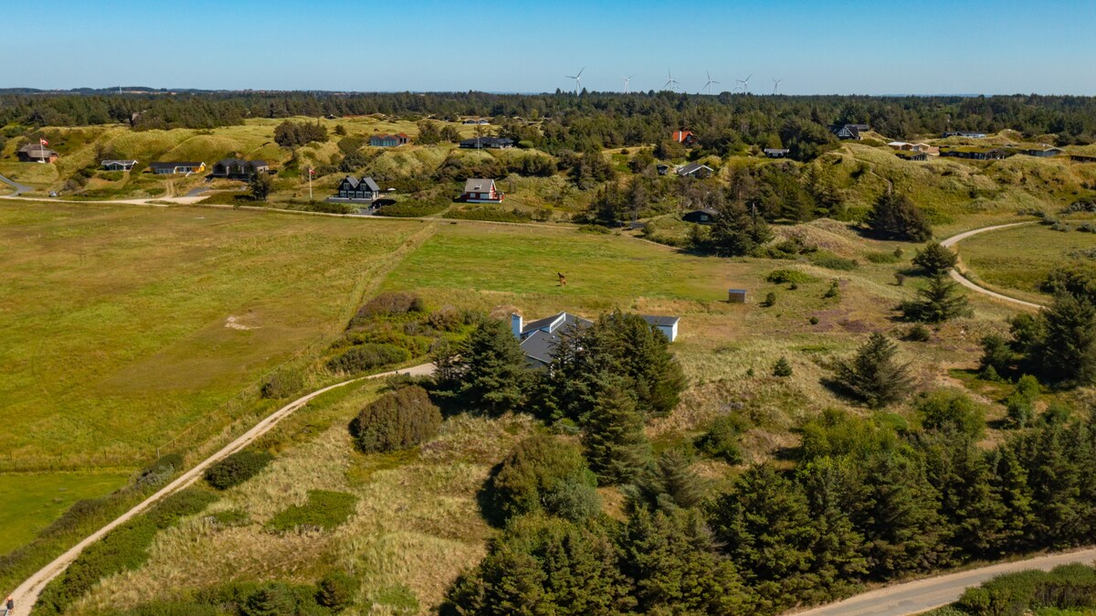 Aerial view capturing a serene landscape characterized by rolling green hills and scattered homes. Wind turbines are visible in the distance. The villa is set amidst greenery, providing a sense of tranquility and space, with natural pathways leading through the countryside.