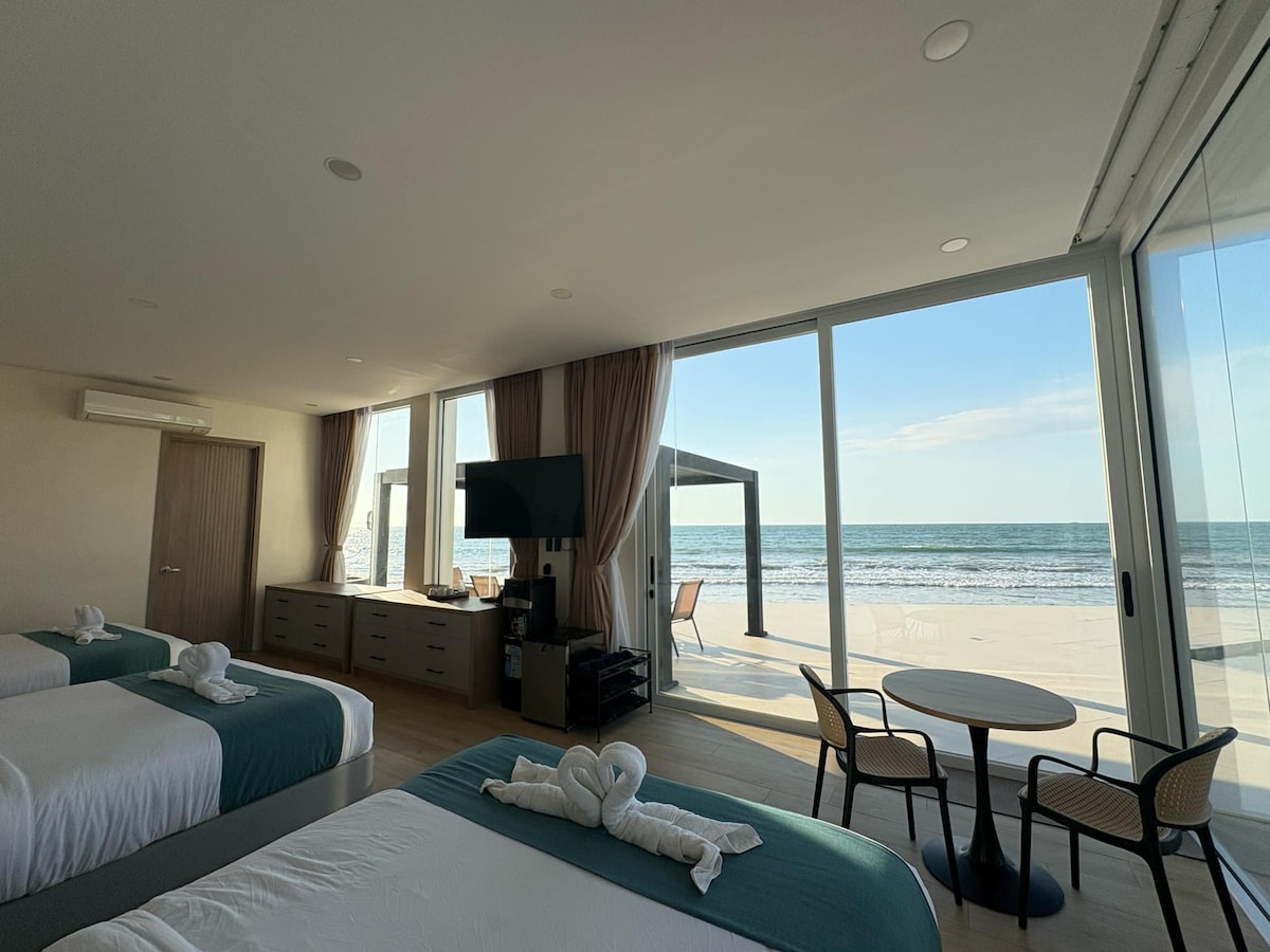 A spacious room with large glass doors offers a clear view of the beach and ocean beyond. Two beds with turquoise accents are complemented by a small table and chairs, inviting relaxation. A flat-screen TV and minimalist furniture enhance functionality.