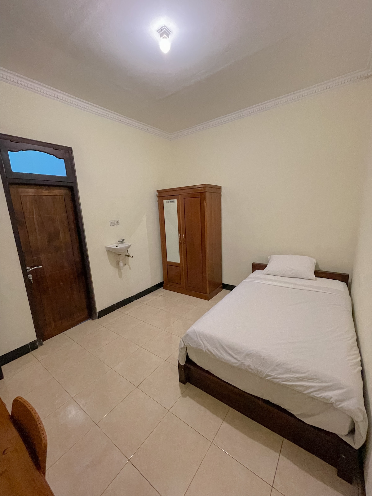 A compact room is equipped with a single bed dressed in crisp white linens. A wooden wardrobe with a mirror stands against the wall, while a small wall-mounted sink adds convenience. Soft lighting illuminates the space, highlighting the neutral color palette.