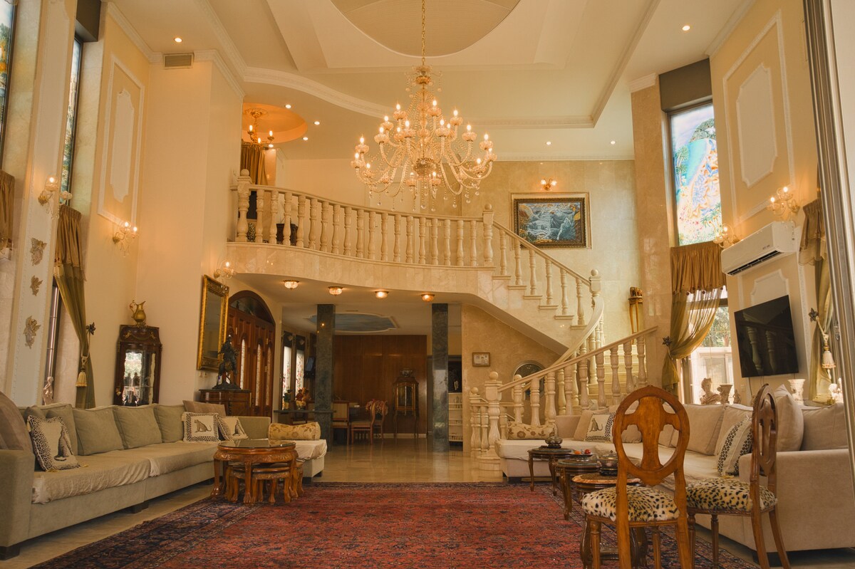An elegantly designed living area features high ceilings and a grand chandelier. The space includes a large, comfortable seating arrangement with plush sofas and ornate wooden accents. A decorative rug lies on the polished floor, leading to a beautifully crafted staircase that enhances the open feel.