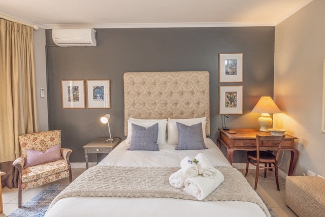 A spacious bedroom is featured, furnished with a Queen size bed covered in crisp linens. Two decorative pillows are placed on the bed. A stylish bedside lamp and a desk with a chair are positioned nearby, alongside a comfortable armchair. Soft wall art and a functional air conditioning unit complement the space.