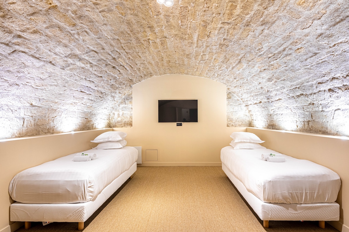 A serene sleeping area is featured, with two single beds positioned on either side of the room. Soft white linens cover the beds, and a television is mounted on the curved stone wall, which is softly illuminated by subtle lighting. A neutral-toned carpet completes the space.