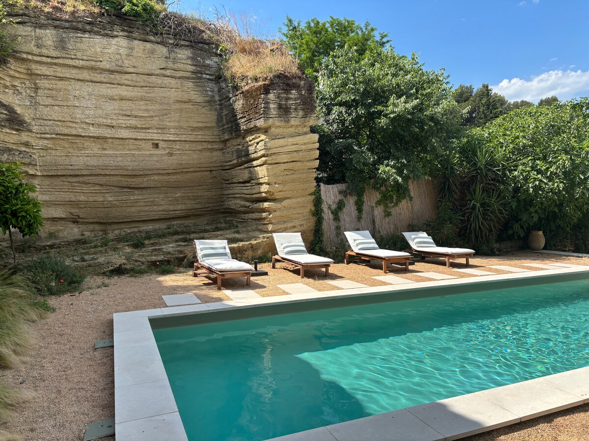 A serene outdoor space features a rectangular swimming pool surrounded by four lounge chairs on a sandy and stone surface. Lush greenery and a natural stone wall provide a calming backdrop, while blue skies are visible above, enhancing the tranquil atmosphere.