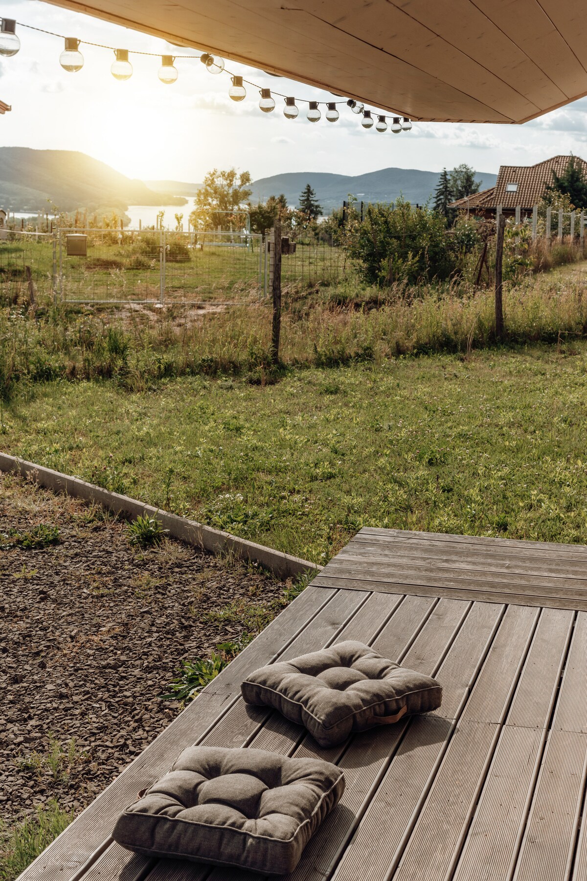 A wooden deck features two cushioned seats, inviting relaxation. The backdrop includes a serene landscape of rolling hills and distant mountains under soft sunset light, while fields and trees create a natural border.