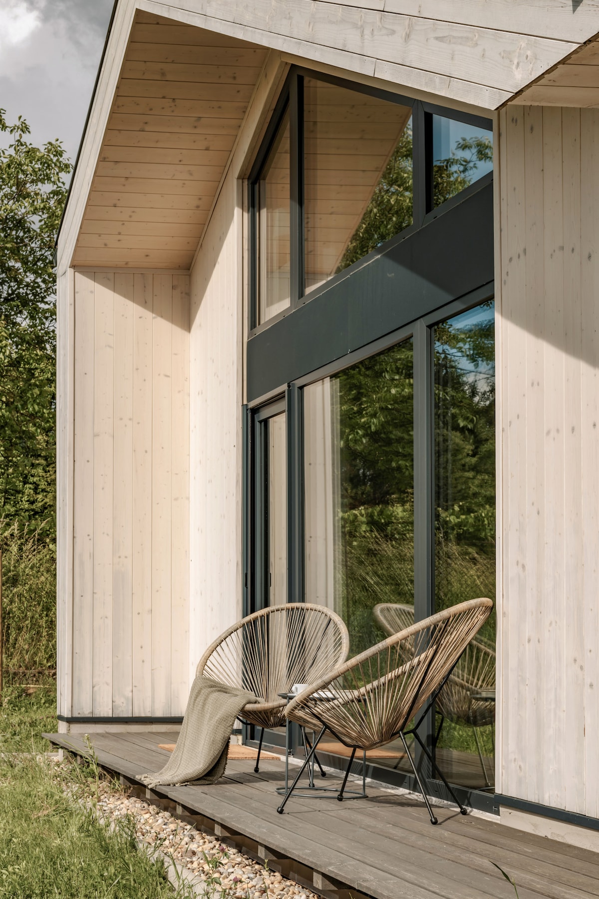 A modern wooden exterior features large glass panels, allowing natural light to flood the interior. Two stylish woven chairs are positioned on a wooden deck, complemented by soft textiles. A lush green landscape surrounds the space, providing a serene outdoor setting.