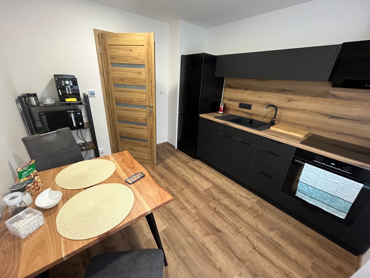 A modern kitchen features sleek black cabinetry and a wooden countertop. A dining table with two placemats is set for two, accompanied by gray upholstered chairs. Kitchen appliances, including a coffee maker and refrigerator, are visible. Warm wooden flooring enhances the contemporary design.