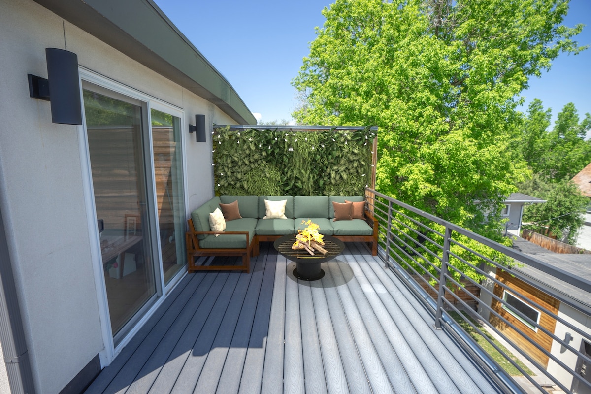 The private roof deck is furnished with a sectional sofa and a round coffee table, surrounded by lush greenery. Large sliding glass doors provide access to the indoor space, while the wooden decking allows for comfortable outdoor seating under clear blue skies.