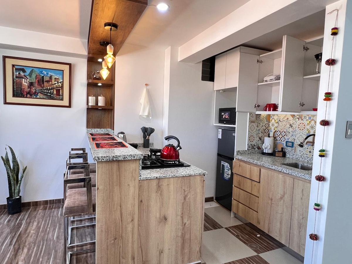 A modern kitchen area features a sleek countertop with bar stools alongside. The cabinetry showcases a combination of wood and white finishes, while the stove and kettle add functional elements. Decorative tiles enhance the vibrant look, complemented by a small green plant in the corner.