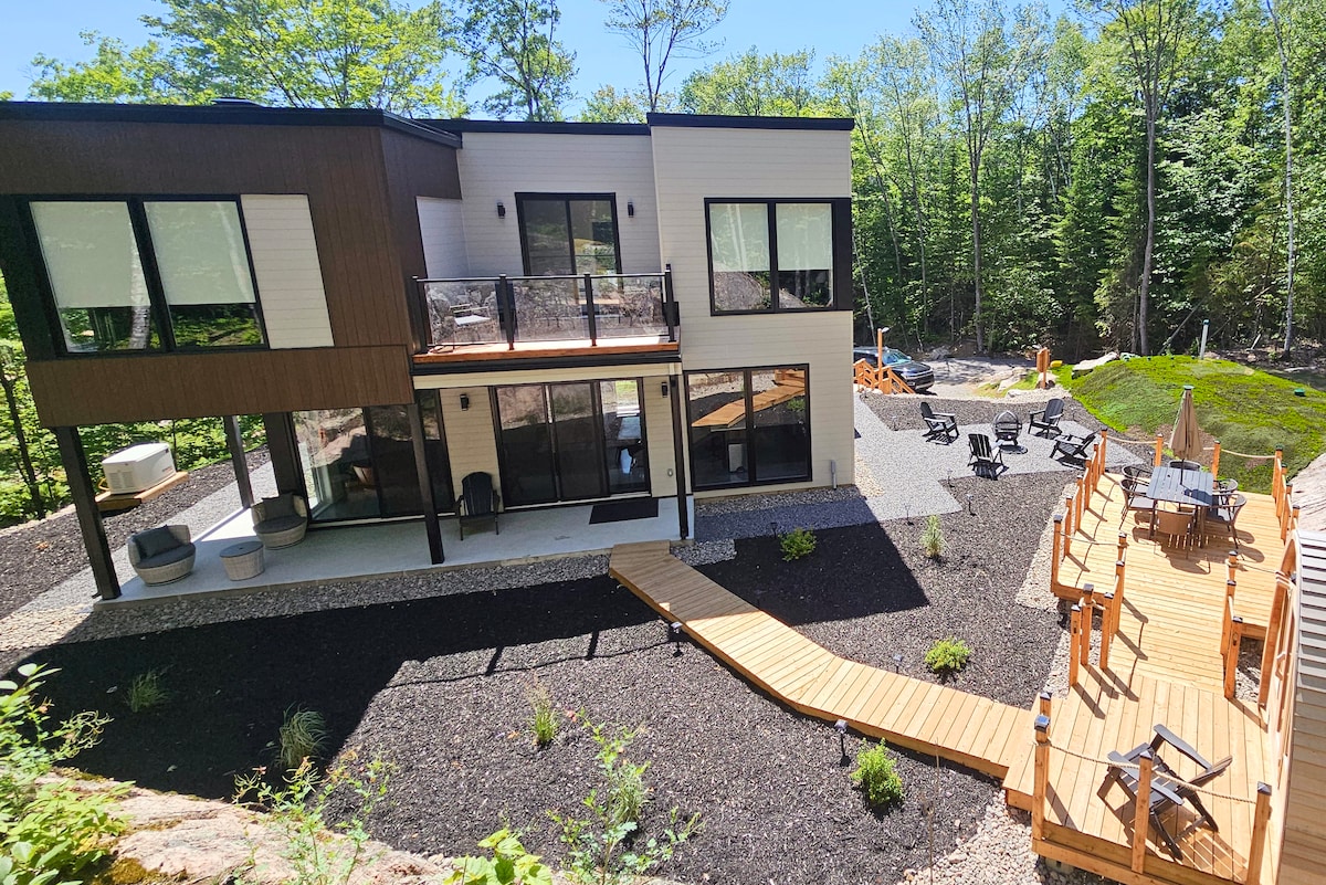 A contemporary chalet is set within a private, tree-lined area. Multiple terraces are seen, including a wooden walkway leading to a patio with seating. The backyard features a fire pit area surrounded by chairs, harmonizing with the natural surroundings.