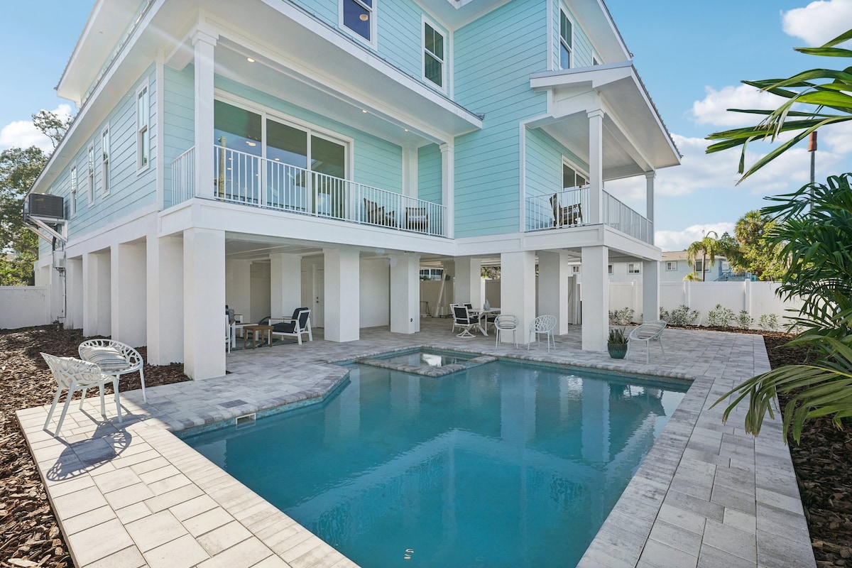 A light blue townhouse features elevated construction with spacious outdoor areas underneath. A rectangular pool is highlighted, surrounded by laid stone tiles. Comfortable seating is arranged nearby, providing a relaxed setting for enjoying the poolside.