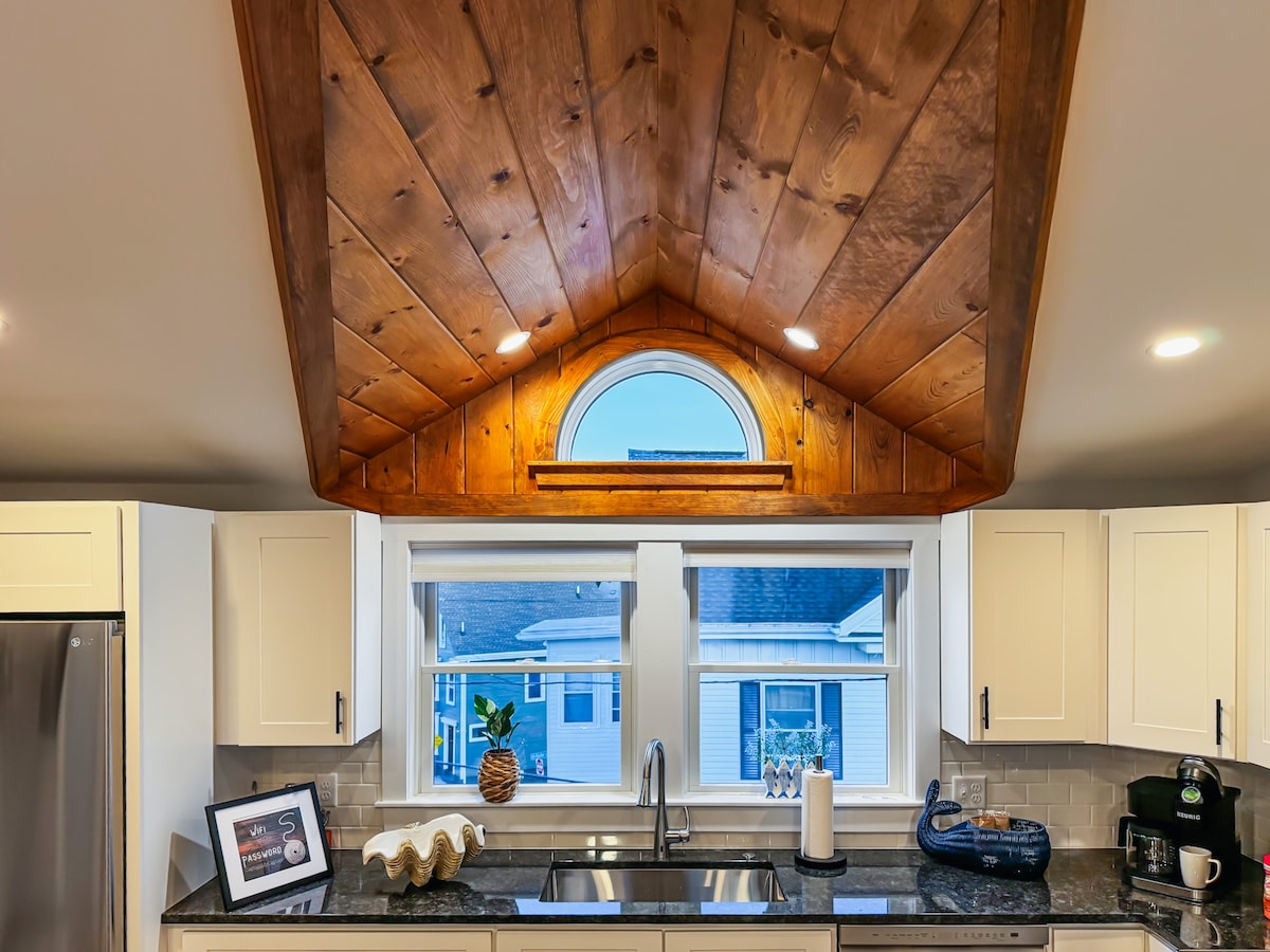 A bright kitchen features a warm wood-paneled ceiling with arched windows that allow natural light to fill the space. White cabinetry contrasts with dark countertops. Decorative elements, including a shell and a framed photo, are placed on the counter, enhancing the home's welcoming design.