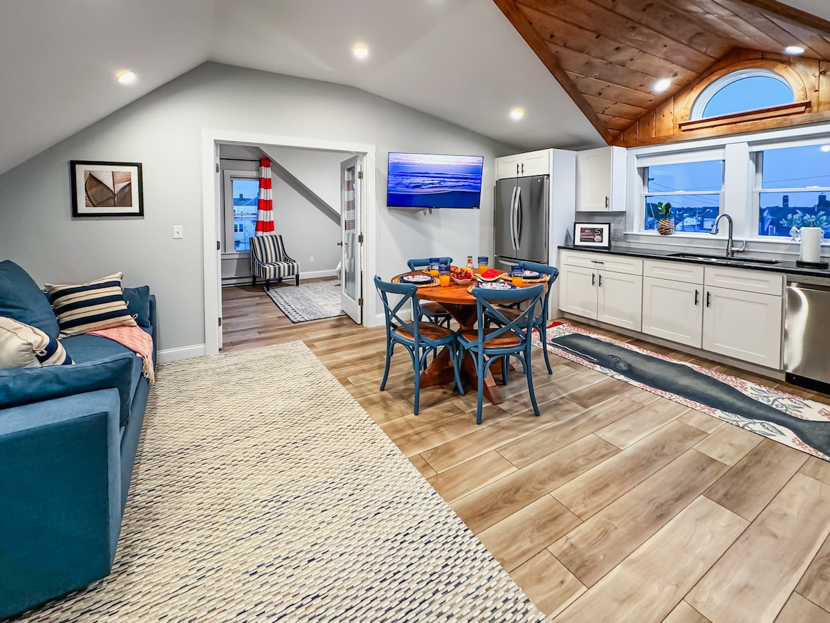 A contemporary open-plan living area is showcased, featuring a blue couch, a round dining table set for four, and modern kitchen appliances. Natural light fills the space through large windows, and sloped wooden ceilings add a cozy touch to the atmosphere.