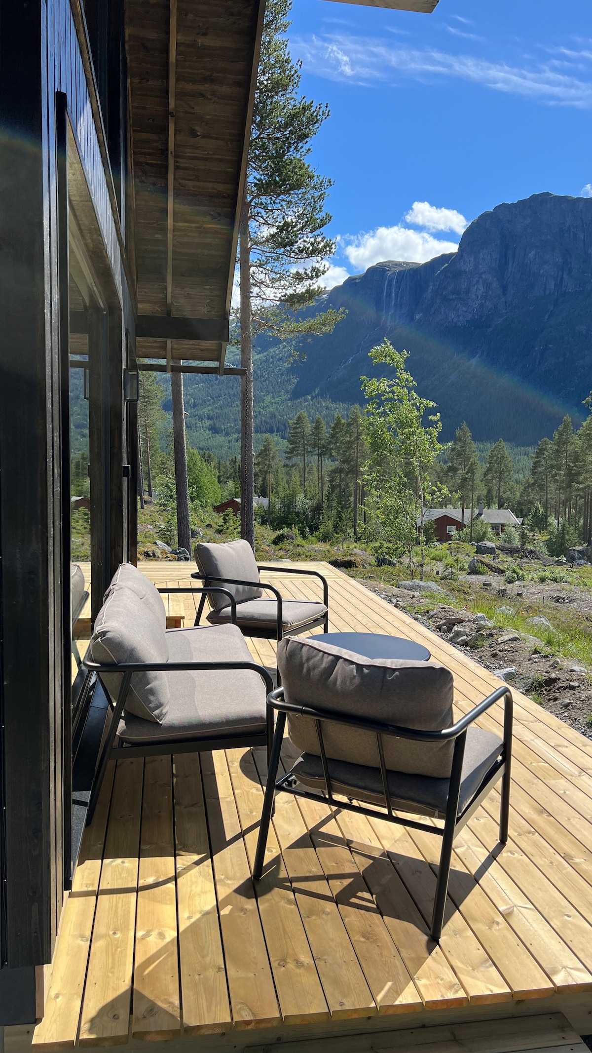 A spacious wooden deck features two modern chairs, positioned to offer views of the surrounding landscape. Lush greenery and mountains can be seen in the background, with sunlight filtering through the clouds, creating a serene outdoor space for relaxation.