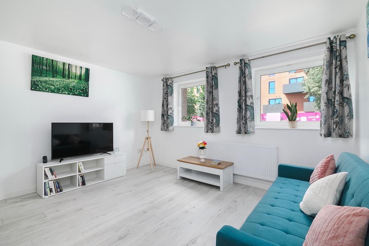 A bright living area features a teal couch and a coffee table placed on light-colored flooring. A television is positioned against one wall, alongside a shelf containing books. Natural light enters through two windows adorned with patterned curtains, enhancing the space's openness.