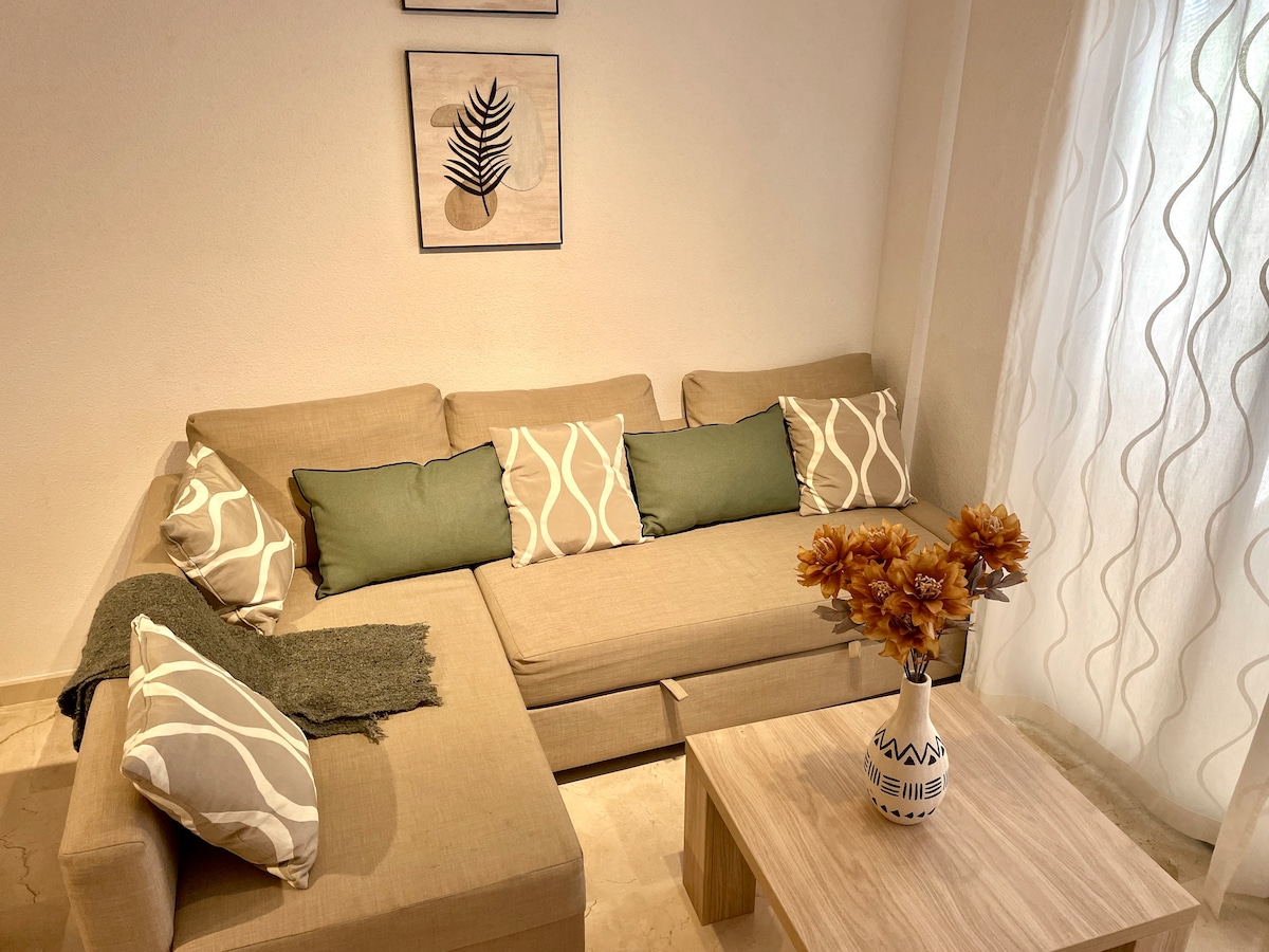 A spacious seating area features a contemporary sectional sofa adorned with decorative cushions in neutral tones. A light wooden coffee table sits in the center, complemented by a vase of artificial flowers. Natural light filters through sheer curtains, creating a bright and inviting space.