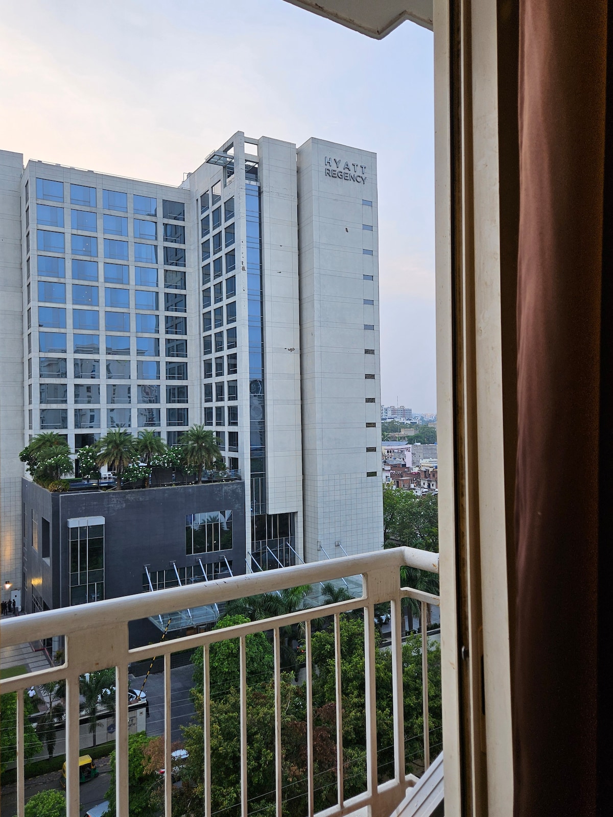 A view from a balcony captures a modern hotel building with large glass windows. The surrounding area features greenery and urban architecture, providing a sense of the city landscape. Light can be seen reflecting off the building, enhancing the scene.