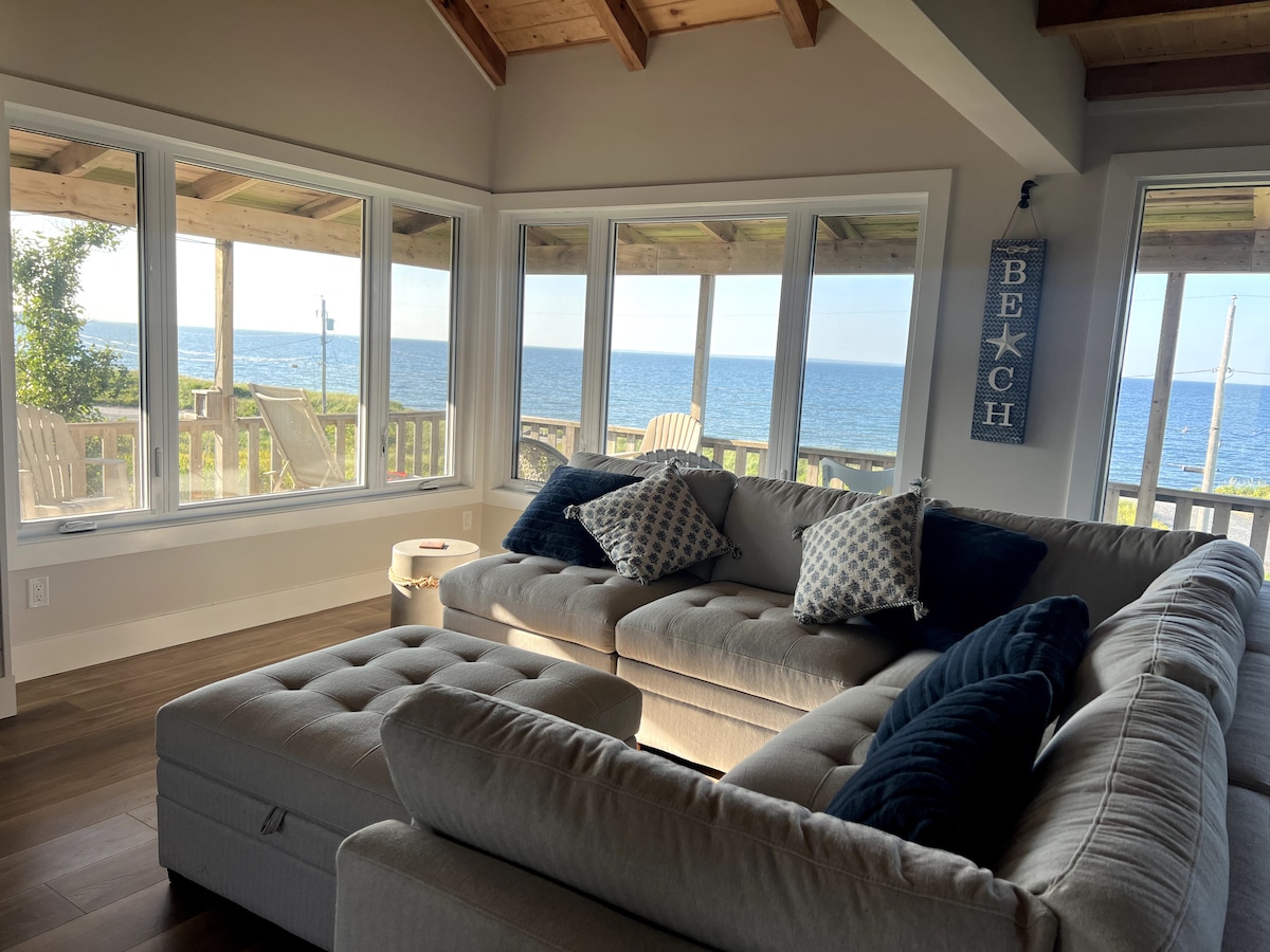 A spacious living area features a large sectional sofa with decorative pillows, positioned near expansive windows that offer a panoramic view of the ocean. Natural light fills the room, accentuating the wooden beams of the ceiling and the inviting seating arrangement.