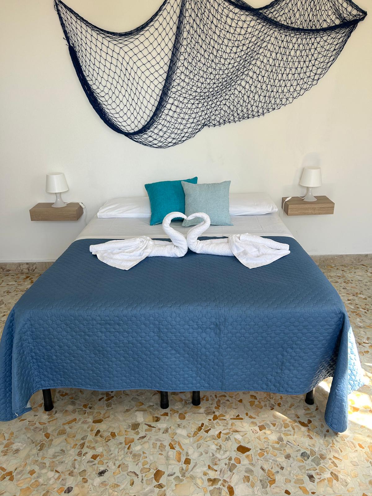 A spacious bed is dressed in a blue quilt, complemented by decorative cushions in shades of turquoise. Two swan-shaped towels are elegantly arranged on the bed. Simple lamps are positioned on the nightstands, with a decorative net hanging above, creating a serene ambiance.