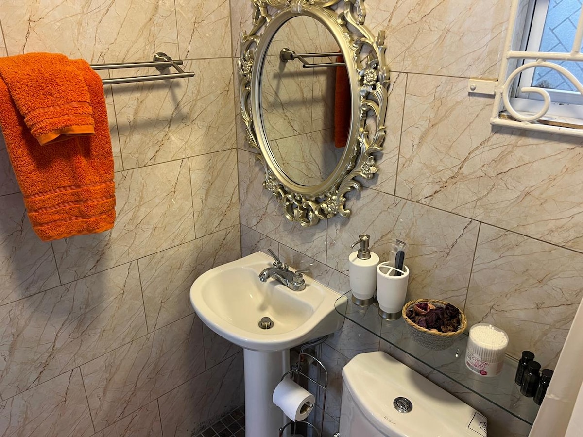 The bathroom features elegant tile work and a decorative round mirror framed in ornate gold. A white pedestal sink is complemented by a set of neatly arranged toiletries on a glass shelf, while bright orange towels add a pop of color to the space.