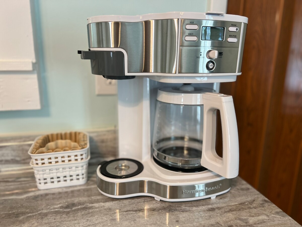 A modern coffee maker with stainless steel accents is positioned on a smooth granite countertop. It features a glass carafe resting on a hot plate, with a digital display and control buttons. Nearby, a white basket holds coffee filters for convenience.