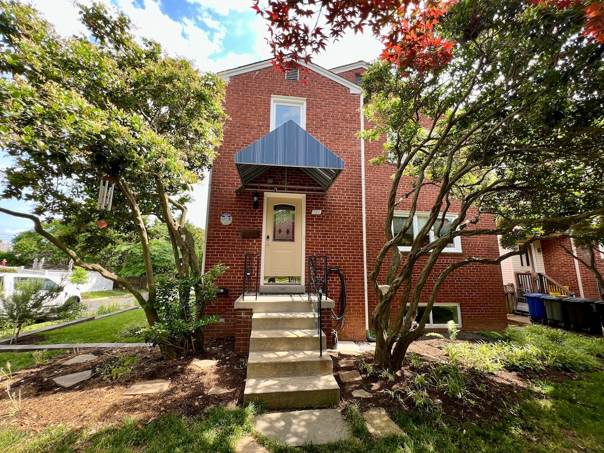 A welcoming exterior view of the brick building, featuring a concrete stairway leading to a door under a blue canopy. Surrounding foliage includes lush green trees and shrubs, enhancing the inviting entrance. A glimpse of outdoor space can be seen to the left.