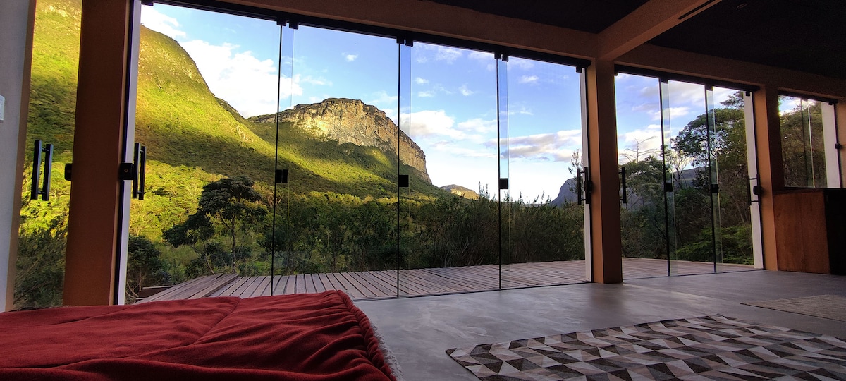 Floor-to-ceiling glass doors open to a stunning view of rolling hills and mountains. A wooden deck extends outside, surrounded by greenery, while natural light fills the interior space featuring a cozy seating area with a warm blanket draped over the seating.