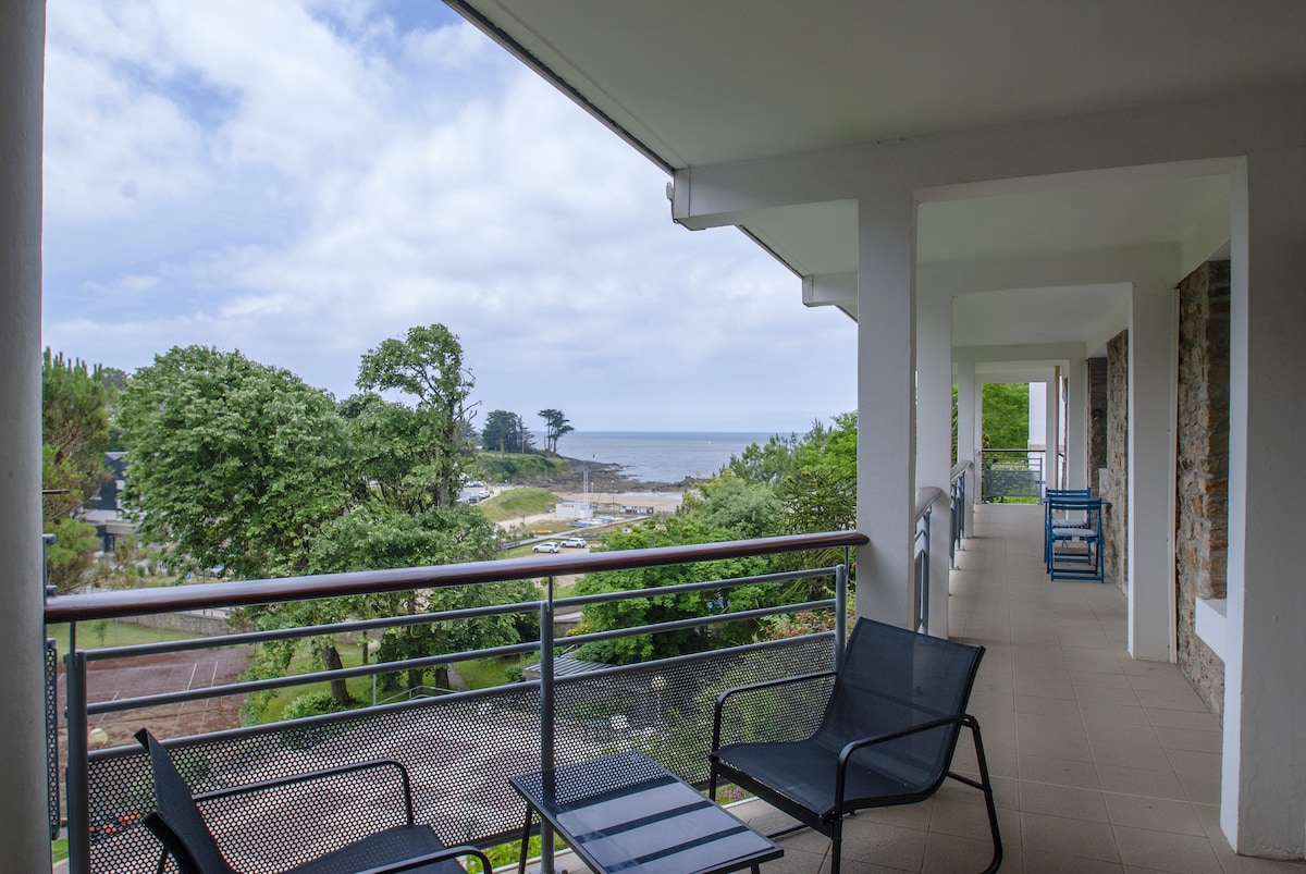 An outdoor terrace is visible, featuring two black lounge chairs. A view of lush greenery and the ocean is provided in the background, with a cloudy sky above. The space is designed for relaxation, inviting guests to enjoy the coastal scenery.