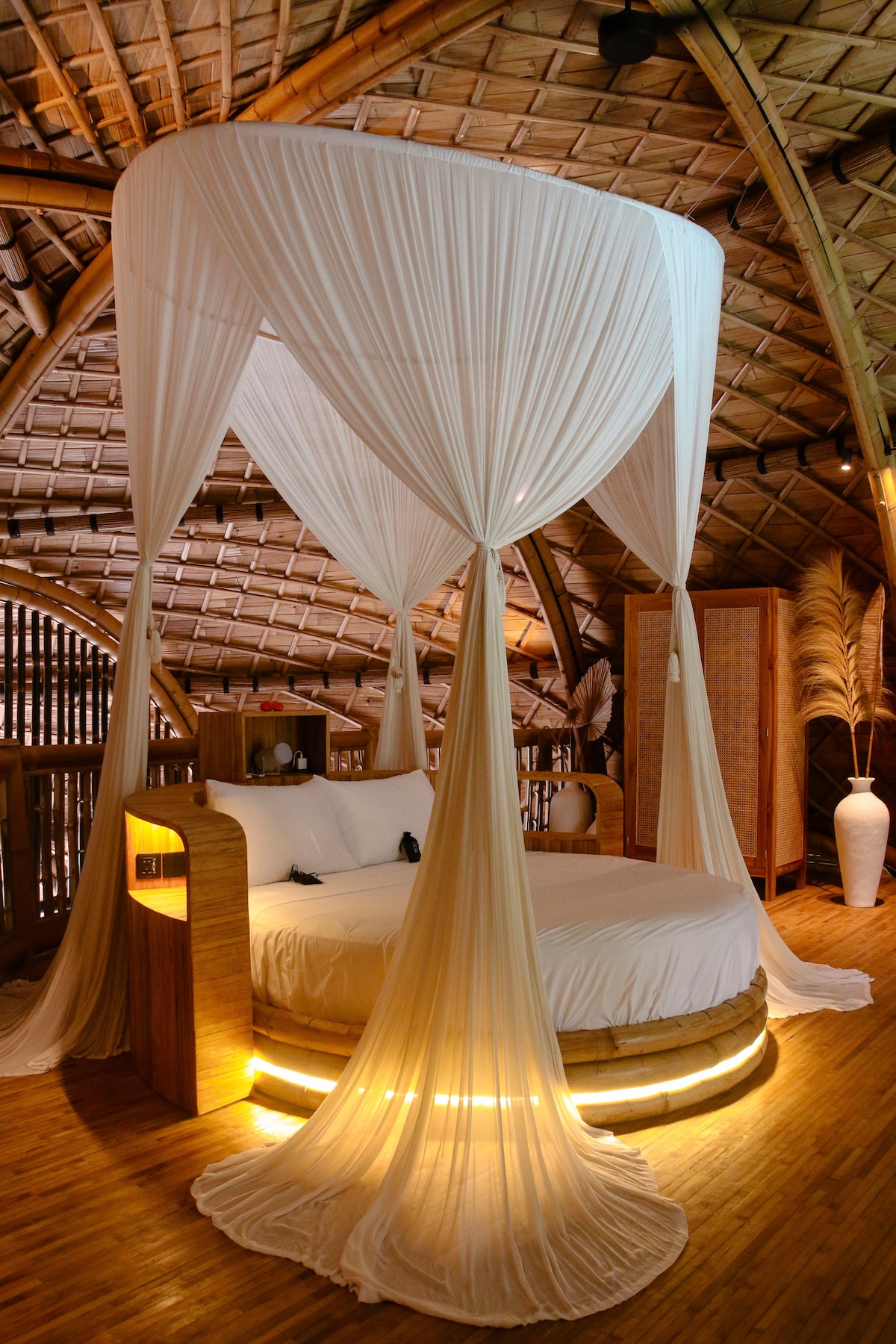 A spacious bed is surrounded by flowing, sheer drapery, creating a serene sleeping environment. The bed's base features illuminated accents, enhancing the overall warmth of the room. The bamboo ceiling showcases the villa's natural architecture, providing a rustic yet elegant aesthetic.