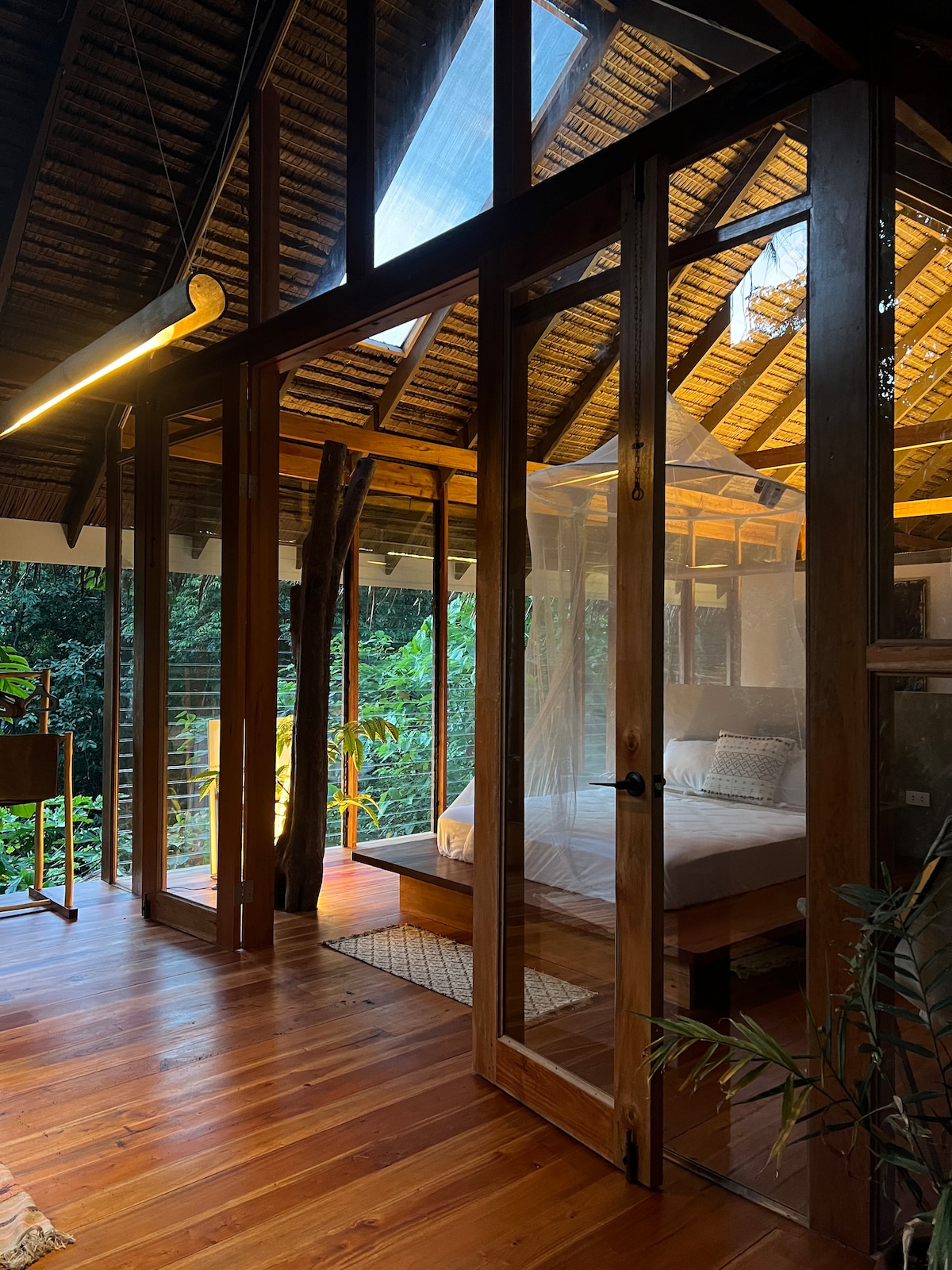 A spacious interior showcases a cozy sleeping area with a large bed, illuminated by natural light coming through floor-to-ceiling glass doors. The warm wooden flooring complements the rustic ceiling, while views of the surrounding greenery create a serene atmosphere.