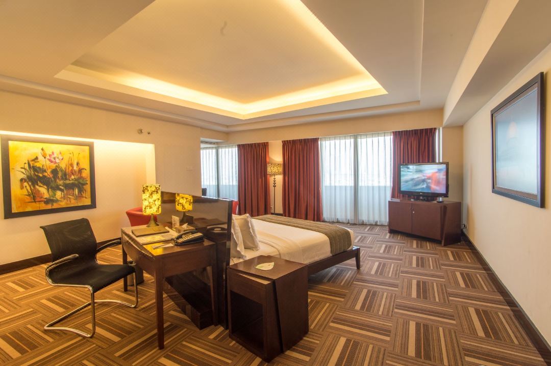 A spacious room features a plush bed positioned against a wall with soft lighting. A work desk and chair are situated nearby, while a flat-screen TV is supported on a wooden cabinet. Large windows with flowing curtains allow ample natural light.