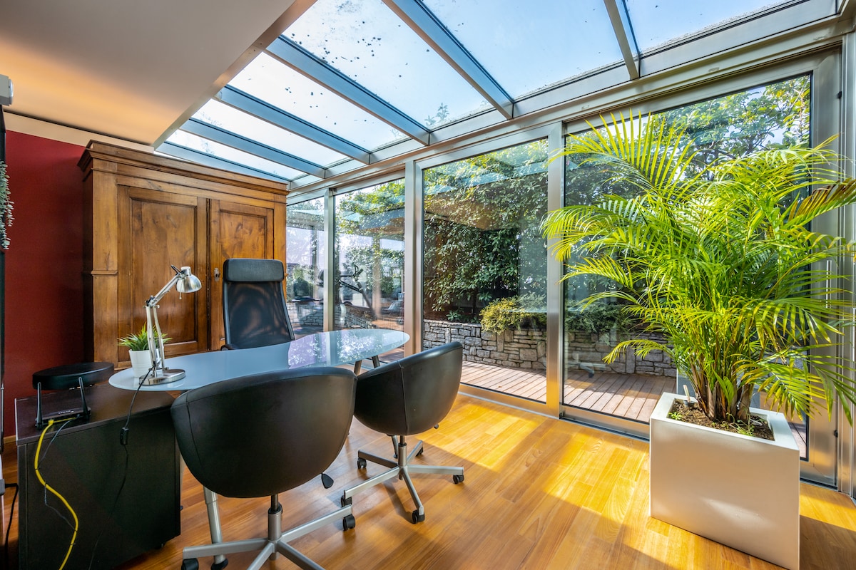 A sunlit workspace features a modern, glass-enclosed design with wooden flooring. A sleek desk is positioned beside a comfortable black chair, and a lush green plant adds a natural touch. Views of a private garden are visible through large glass panels.