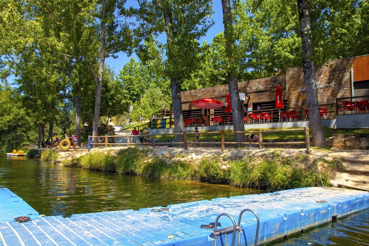 A scenic view of a tranquil river bank is presented, featuring a wooden deck with a swimming ladder. Several trees provide shade, while a casual dining area with red outdoor furniture is visible, inviting relaxation by the water's edge.