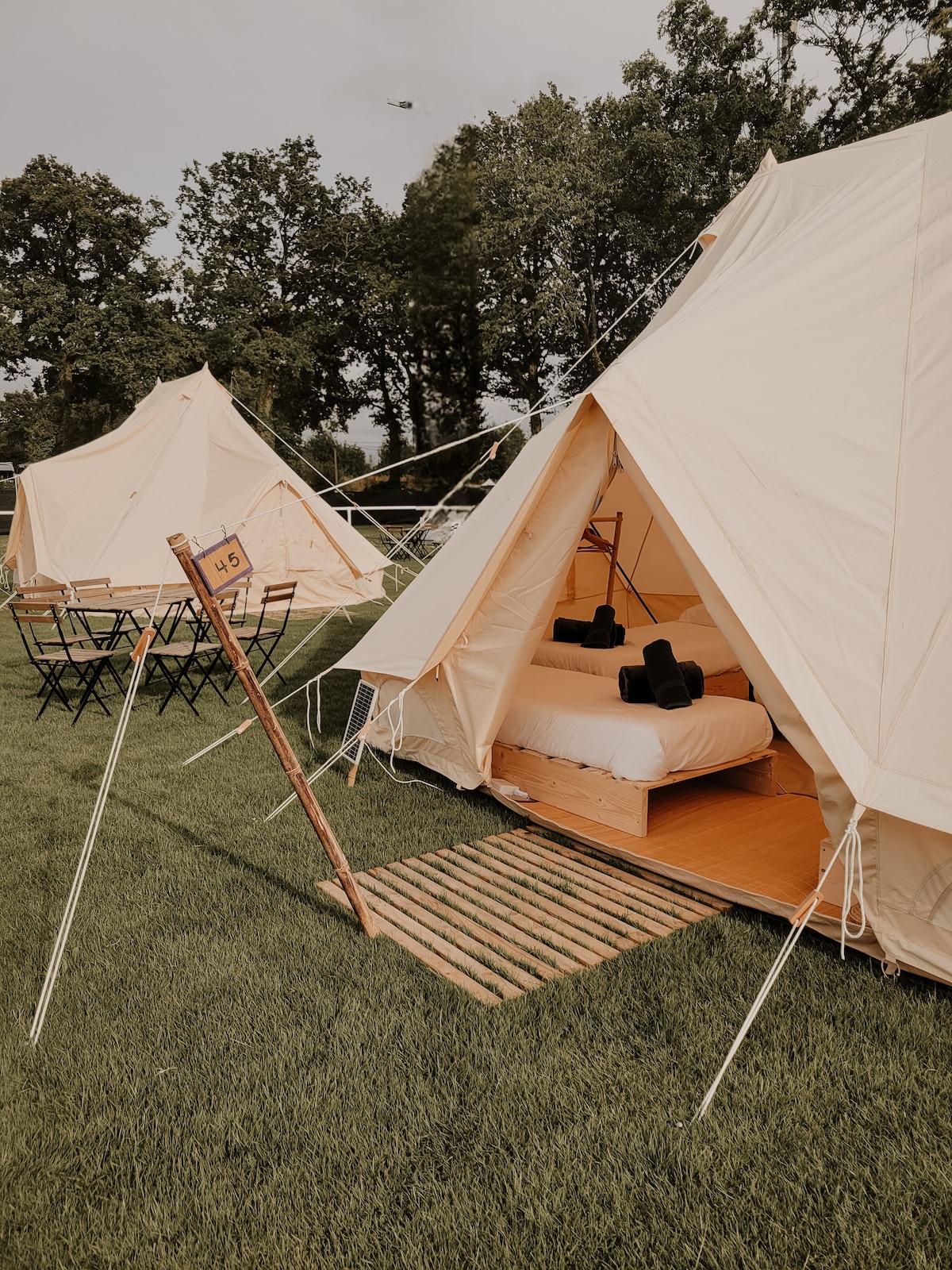 Two bell tents are positioned on a grassy area, each featuring wooden platforms. One tent reveals a cozy sleeping arrangement with a spacious bed and neatly placed cushions. Nearby, a wooden sign indicates the tent number, while folding chairs are arranged outside for seating.