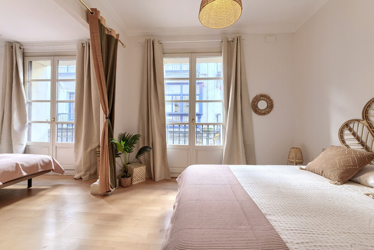 A bright sleeping area features large windows with light filtering through sheer curtains. A spacious bed is adorned with neutral-toned linens, while a separate bed is positioned nearby. Decorative elements include a rattan wall hanging and a potted plant, enhancing the airy feel of the room.