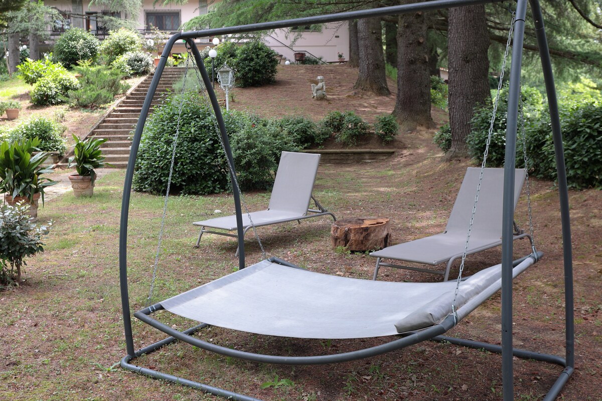 A serene outdoor space features a garden area, with a swing seat and two lounge chairs positioned under the shade of trees. Lush greenery surrounds the seating, creating a peaceful atmosphere for relaxation.
