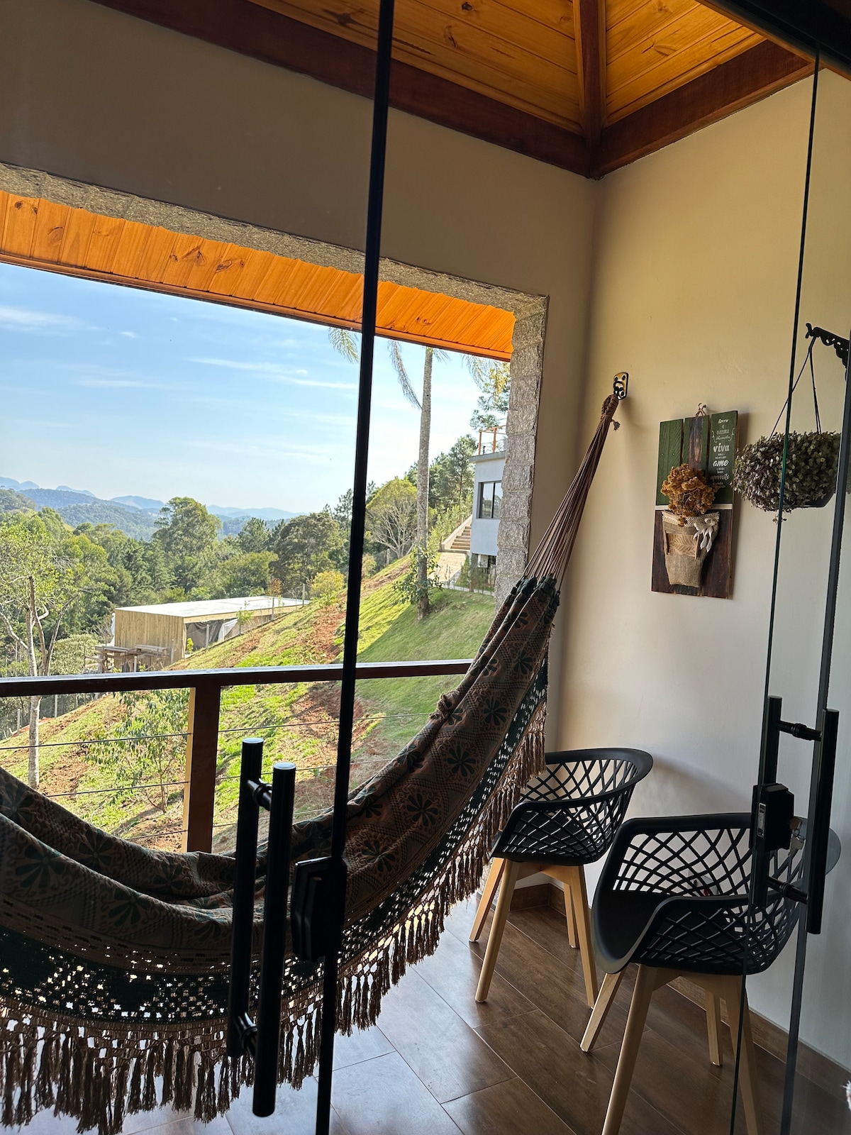 A cozy balcony area features a woven hammock draped between two sturdy supports. Two modern chairs are placed nearby, and a large window showcases a scenic view of rolling hills and greenery. Natural light brightens the space, highlighting the wooden ceiling.
