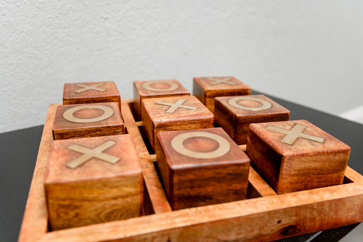 A wooden tic-tac-toe board is presented, featuring nine square blocks arranged in a grid. The blocks are crafted from wood with carved letters 'X' and 'O' on their surfaces, set within a neatly designed wooden frame.