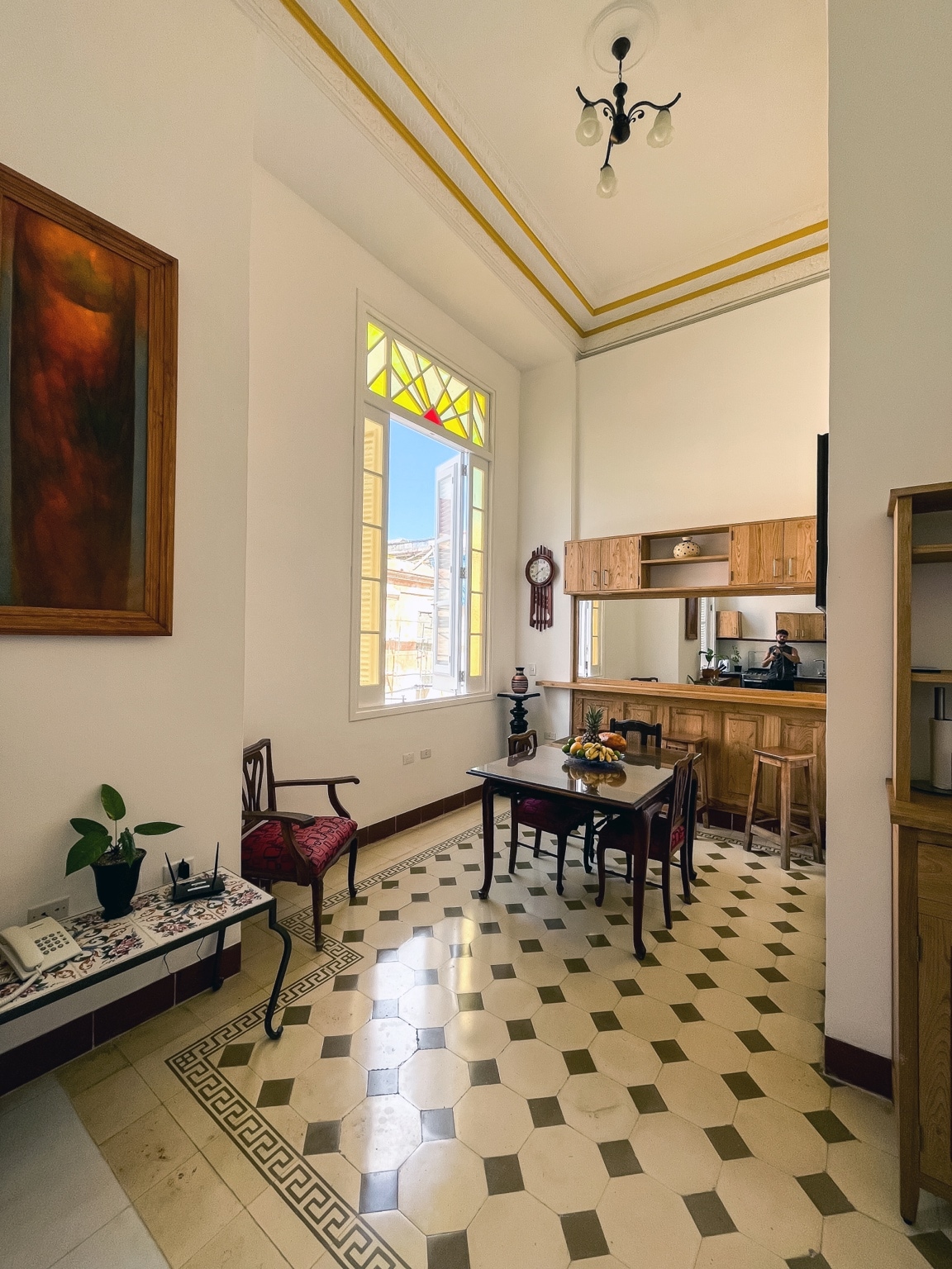A spacious living area features high ceilings and decorative wall accents. A small dining table with chairs is positioned near a colorful stained glass window that allows natural light to brighten the space. The kitchen area, equipped with wooden cabinetry, is visible in the background.