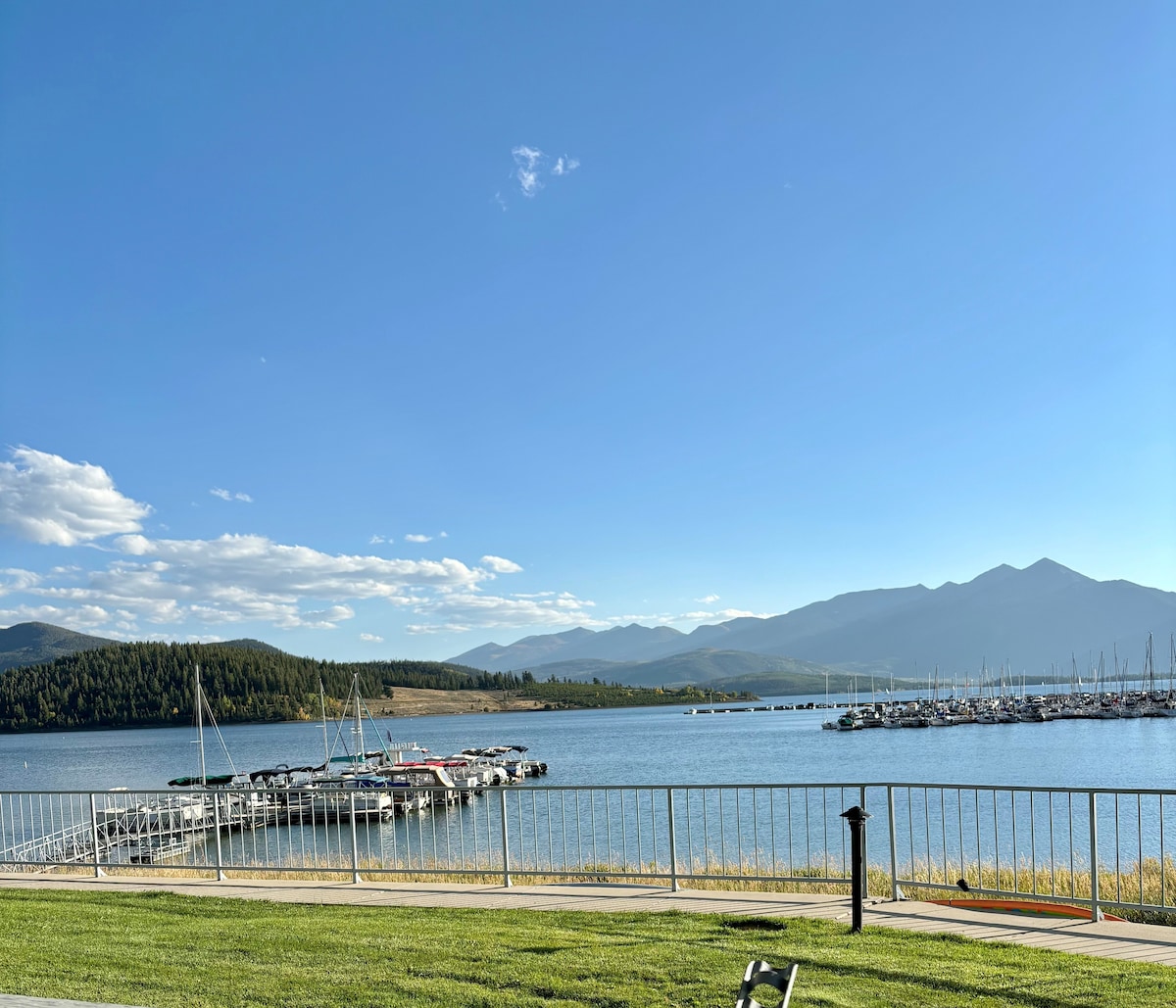 A clear view of Lake Dillon extends towards the horizon, framed by distant mountains under a bright blue sky. The scene includes a marina with several boats docked along the waterfront, while lush greenery is visible in the foreground.