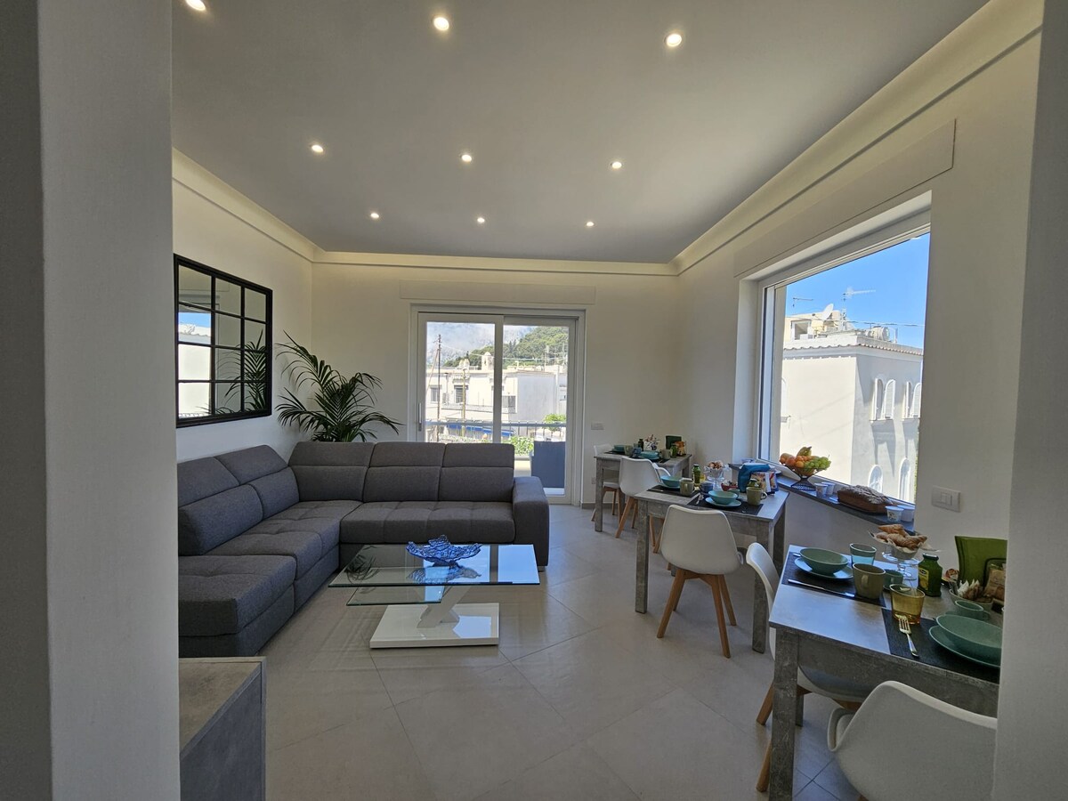 A modern living area is presented, featuring a spacious gray sectional sofa next to a glass coffee table. A pair of large windows allow natural light to flood the space, which is adorned with a dining table set for multiple guests. Decorative plants add a touch of greenery.