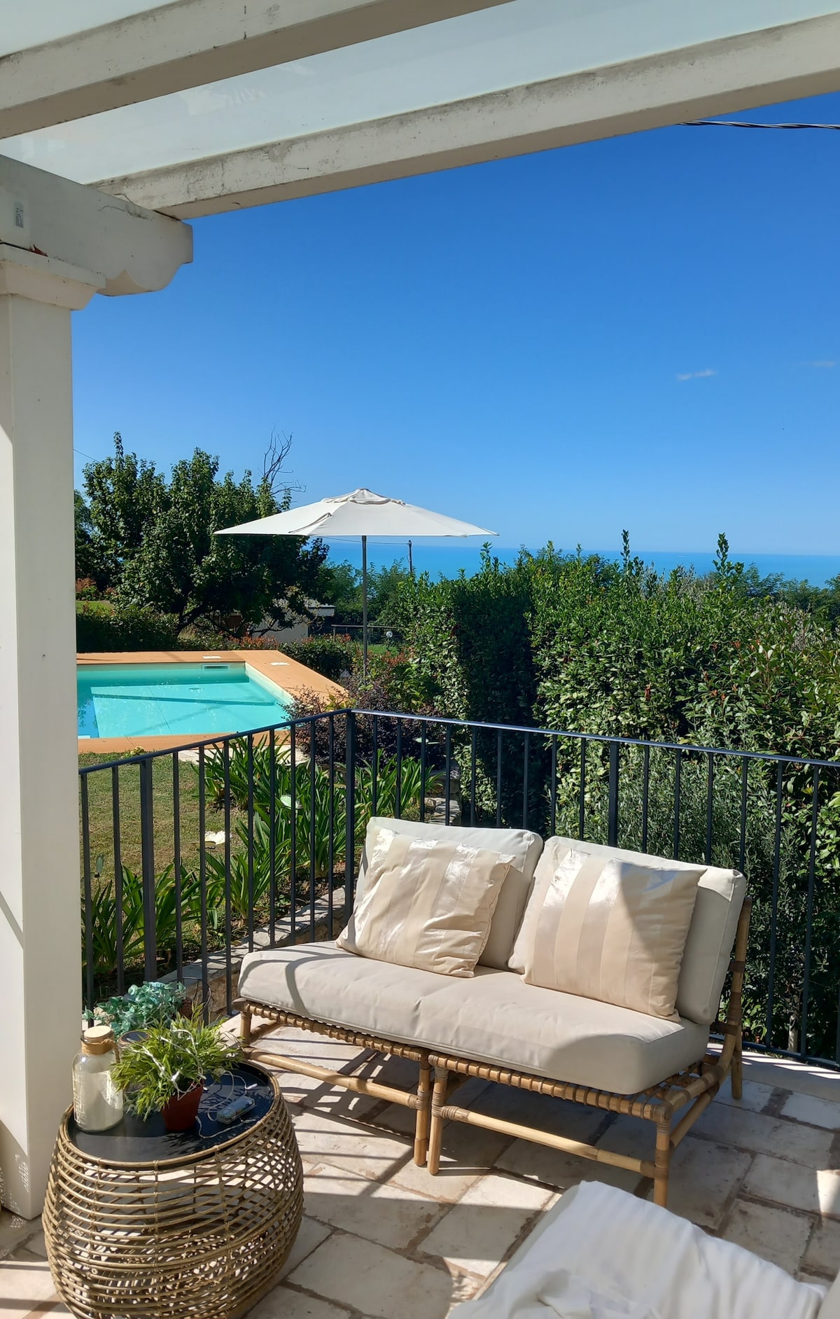 A shaded outdoor seating area features two plush cream-colored cushions on a rattan sofa. In the background, a swimming pool glimmers under the sun, surrounded by lush greenery and a white parasol providing shade. A clear blue sky is visible above.