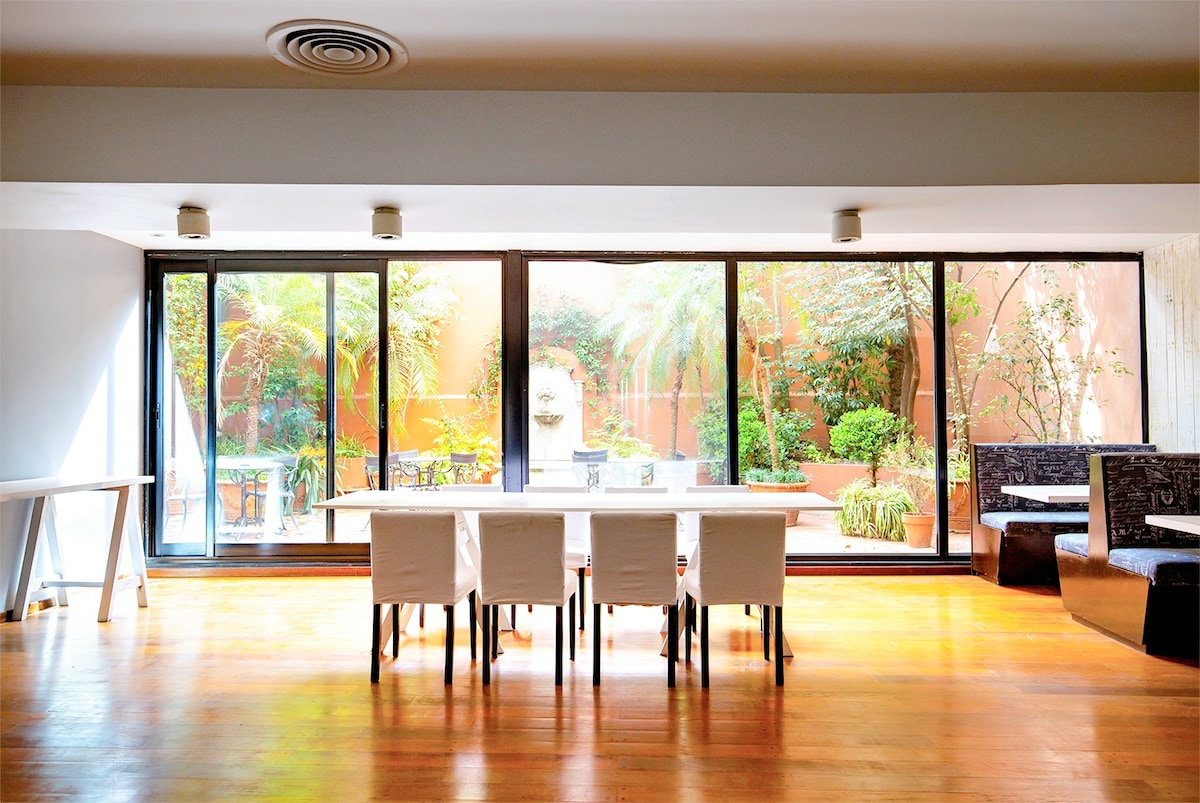 A spacious dining area is displayed with a large table and white chairs, situated in front of expansive glass doors. A lush outdoor garden with tropical plants is visible beyond the glass, creating a bright and inviting space filled with natural light.