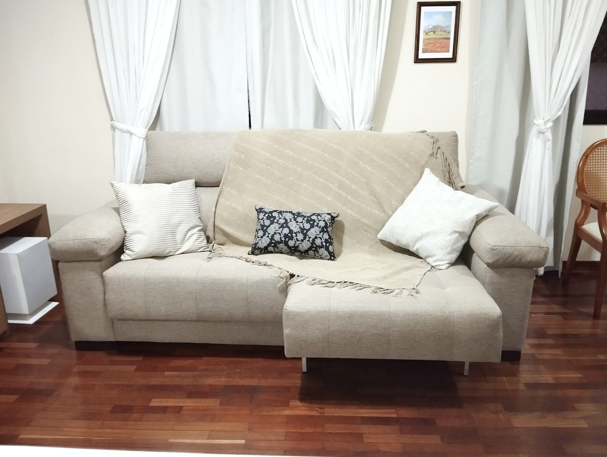 A neutral-toned sofa is positioned against the wall, adorned with a soft throw and decorative pillows. Nearby, sheer white curtains frame a window, allowing subtle light to fill the room. A wooden table and a rattan chair are visible in the background.
