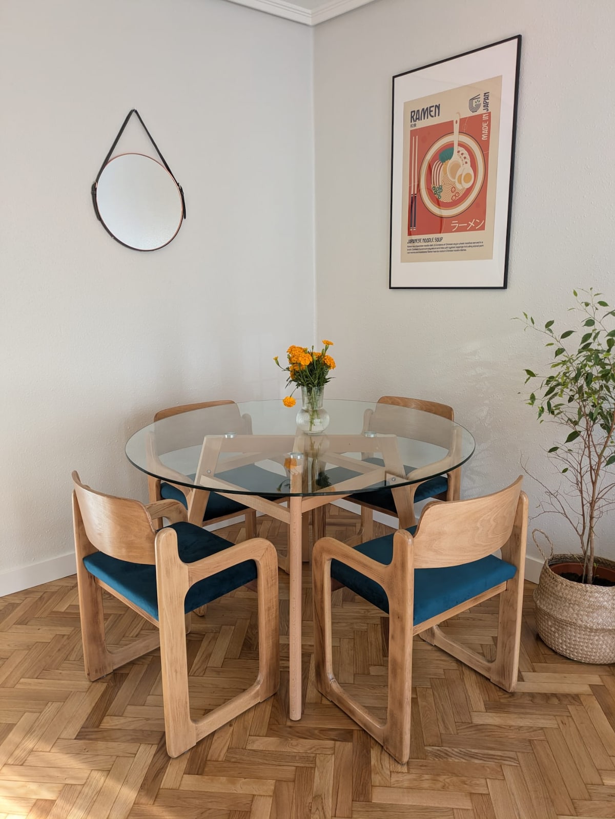 A dining area is presented with a round glass table surrounded by four wooden chairs featuring blue cushions. A vase with fresh flowers is centered on the table. A circular mirror hangs on the wall, complemented by a decorative plant in a basket nearby.