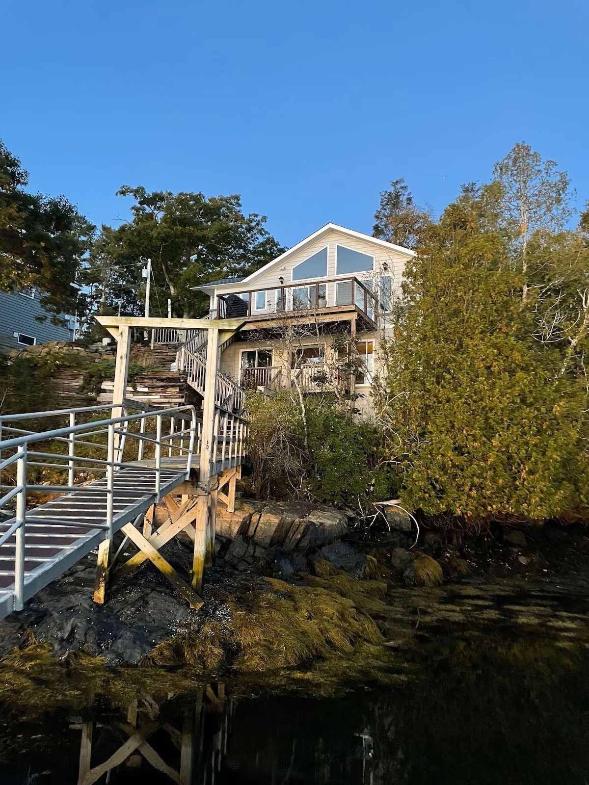 A two-story house sits against a backdrop of greenery, with a wooden dock leading from the water's edge. The structure features decks on both levels, and large windows reflect the evening sky. Surrounding foliage adds to the peaceful ambiance of the location.